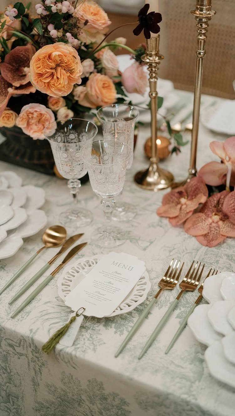 Wedding table setting with peach flowers, gold cutlery, and crystal glasses on patterned linen.