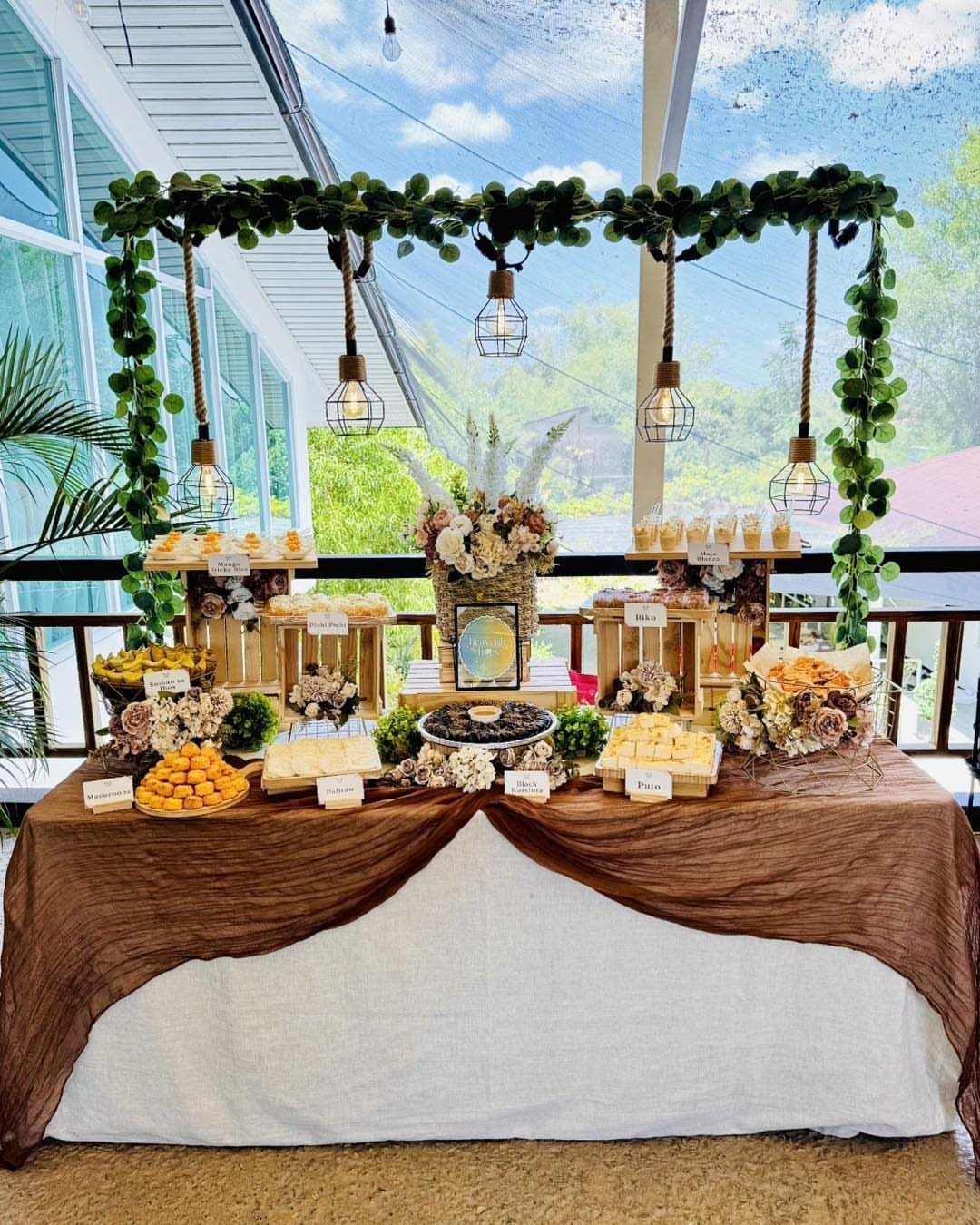 Dessert table with sweets and flowers under a greenery frame with hanging lights