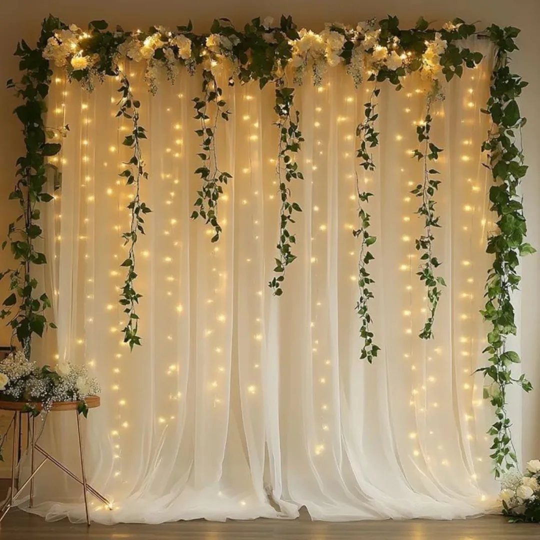 White sheer backdrop with hanging greenery garlands, white flowers, and warm string lights.