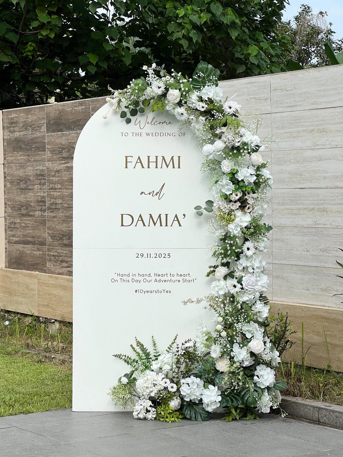 White arched wedding welcome sign with gold text and white floral arrangements.
