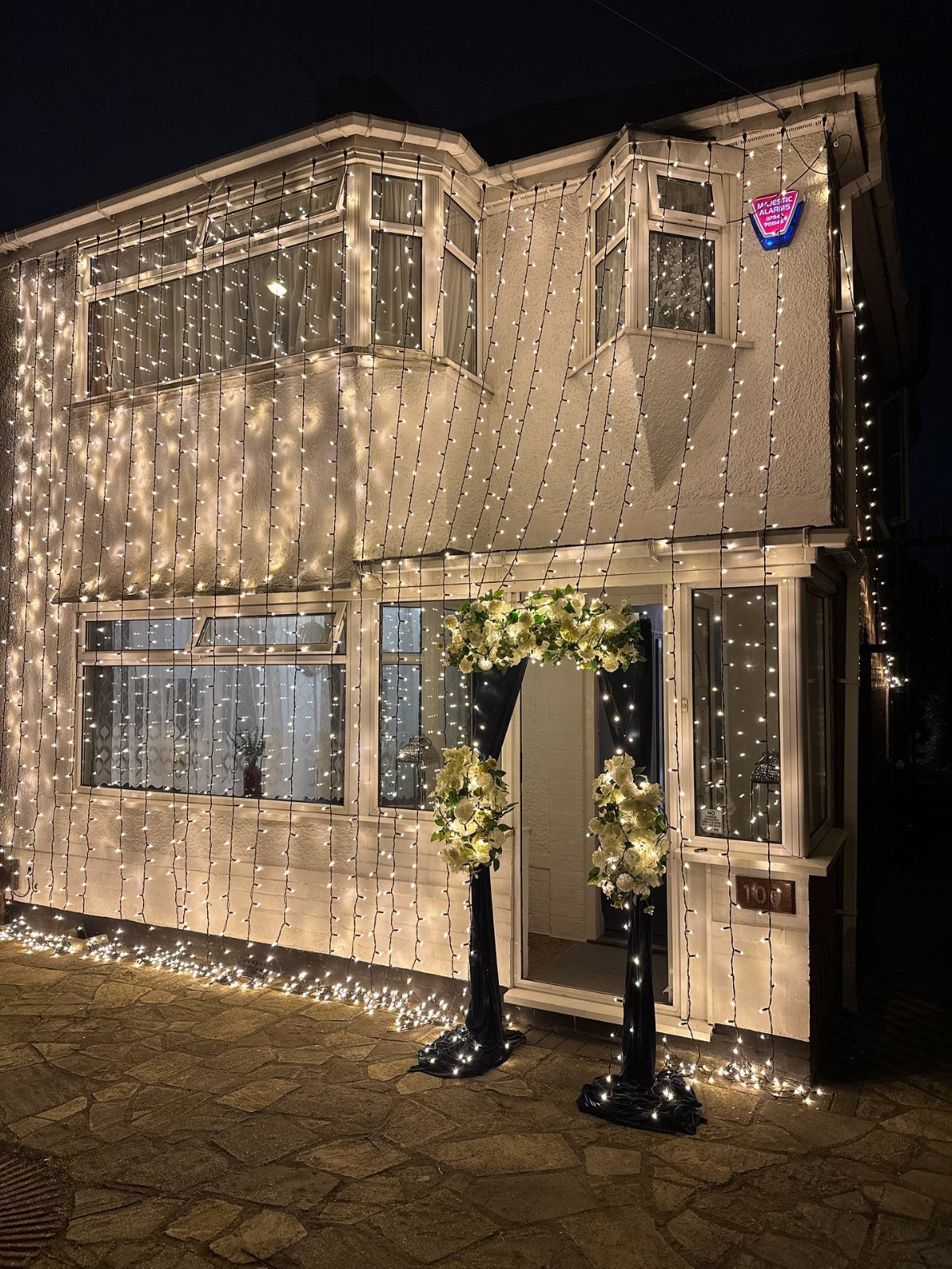 Beige house exterior decorated with cascading warm white string lights and a floral arch.