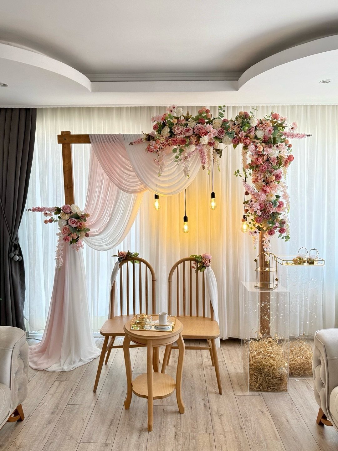 Wooden wedding arch with pink floral arrangements and draped fabric over two chairs.