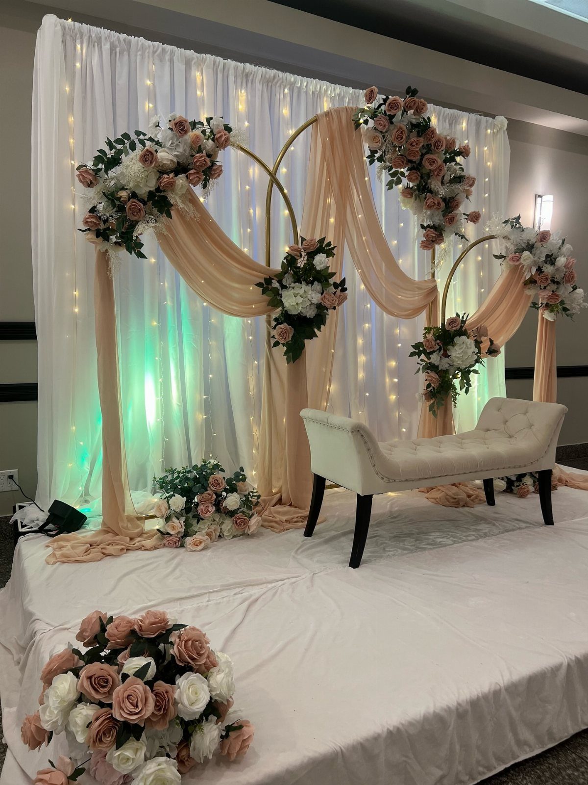 Wedding stage with a white tufted bench, peach fabric, and rose floral arrangements.