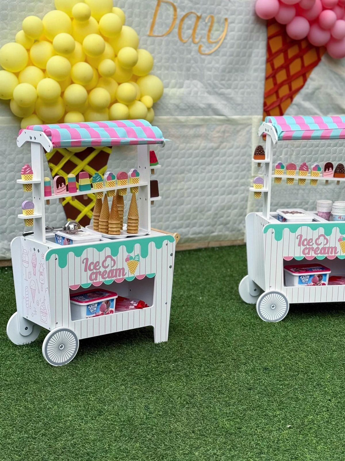 Two white ice cream carts with striped awnings on green grass with balloons.