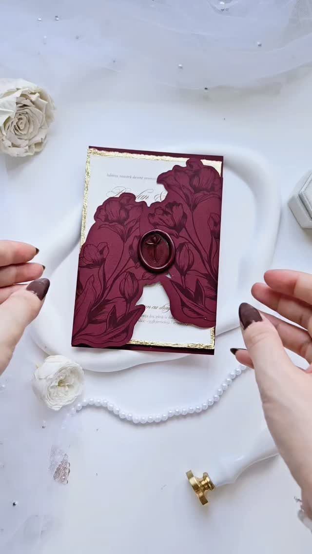 Burgundy laser-cut wedding invitation with floral designs and a wax seal on white tray.