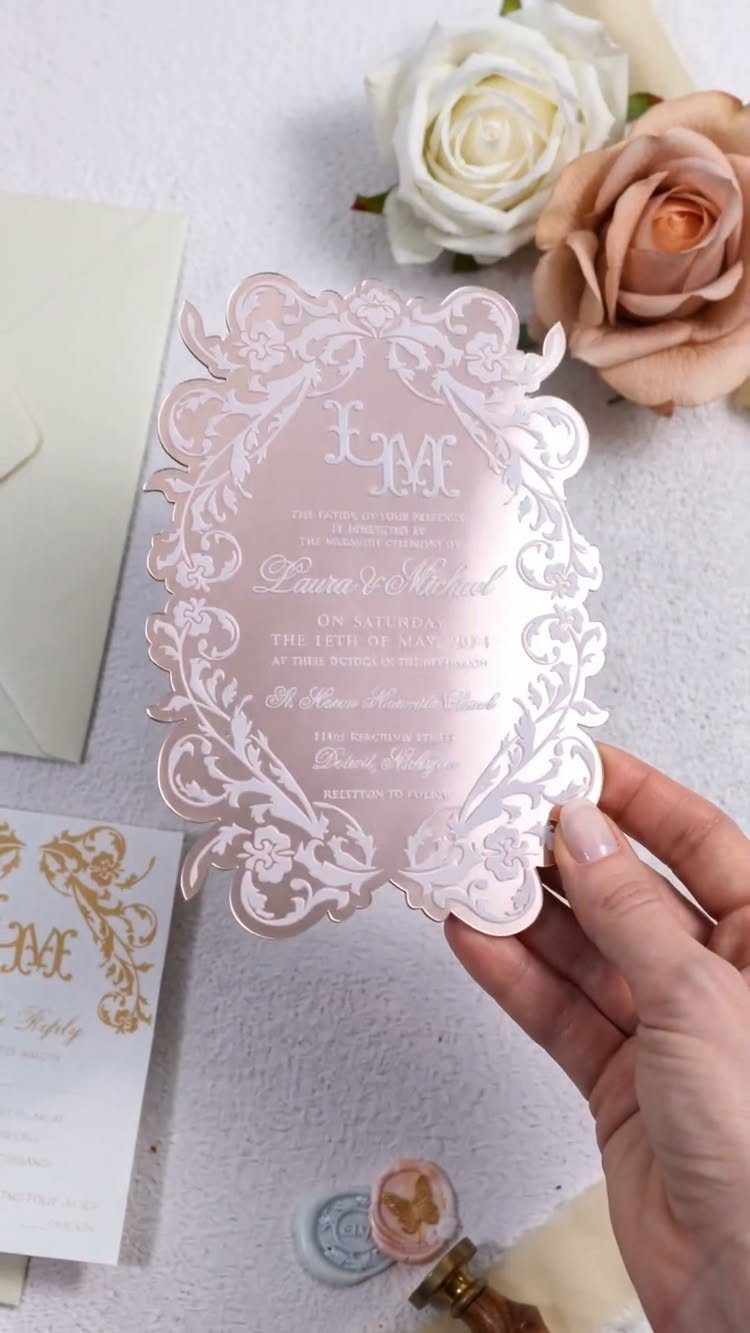 Pink laser-cut wedding invitation with intricate patterns held by a hand near roses.