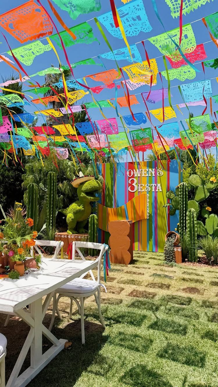 Colorful papel picado banners hanging over an outdoor party area with cacti and dinosaur.