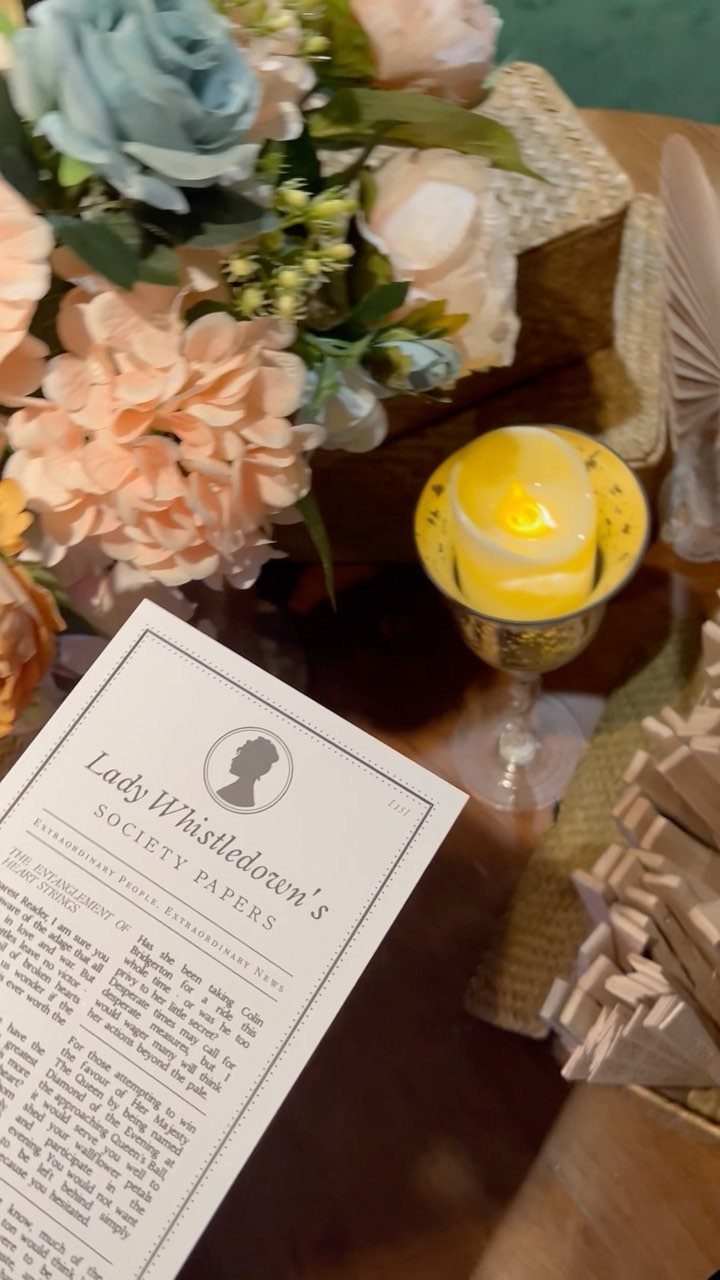 Lady Whistledown's Society Papers next to peach and blue flowers and a lit candle.