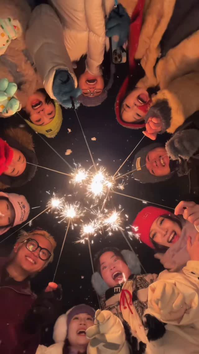 Group of people in winter gear holding lit sparklers in a circle at night