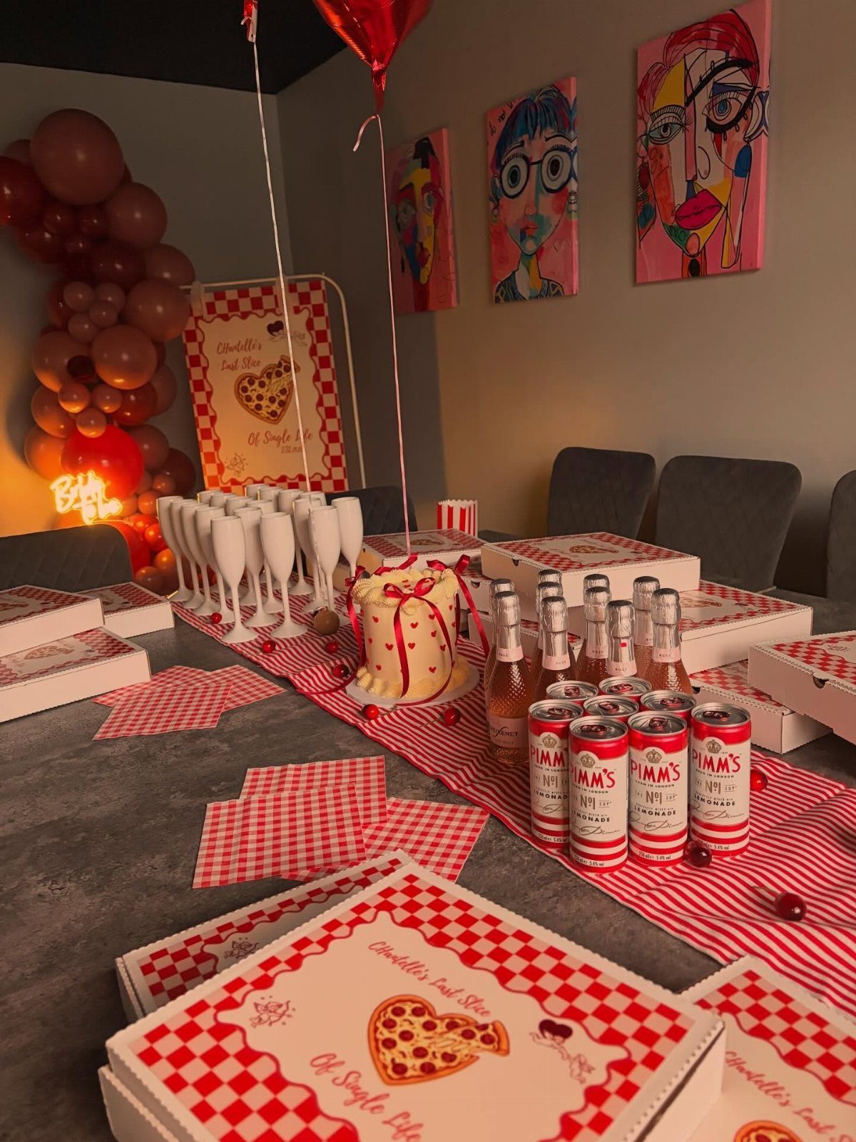 Party table with red checkered linens, pizza boxes, champagne glasses, and a cake.