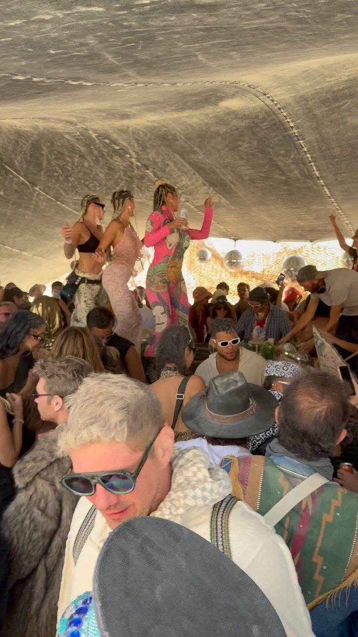 Crowd of adults in bohemian attire dancing and socializing under a large white tent.