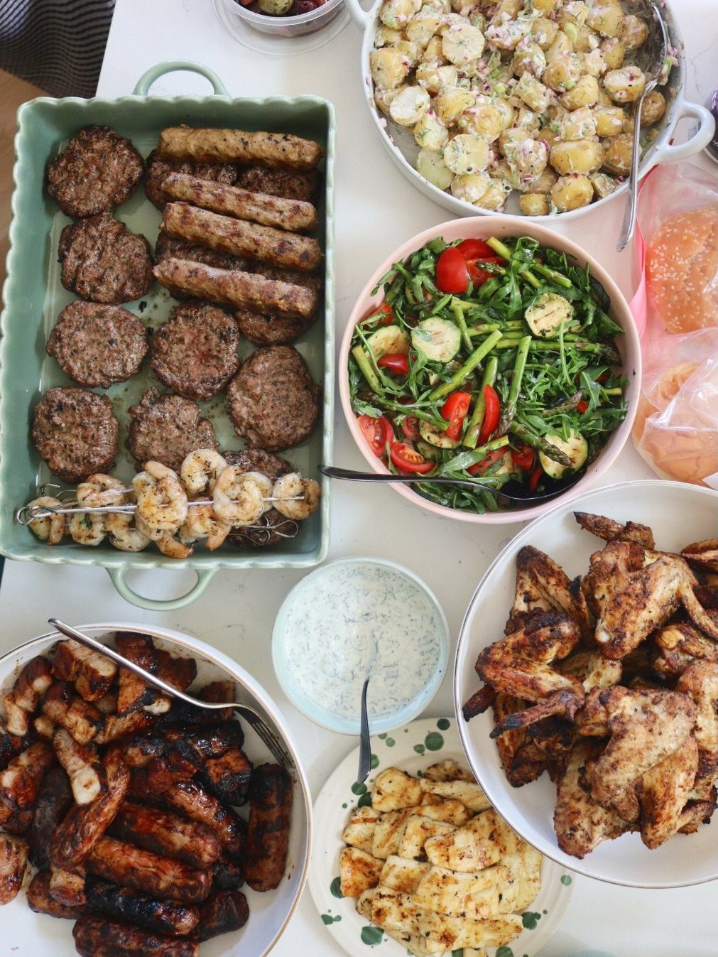 Grilled burgers, sausages, chicken wings, and salads spread across a white table.