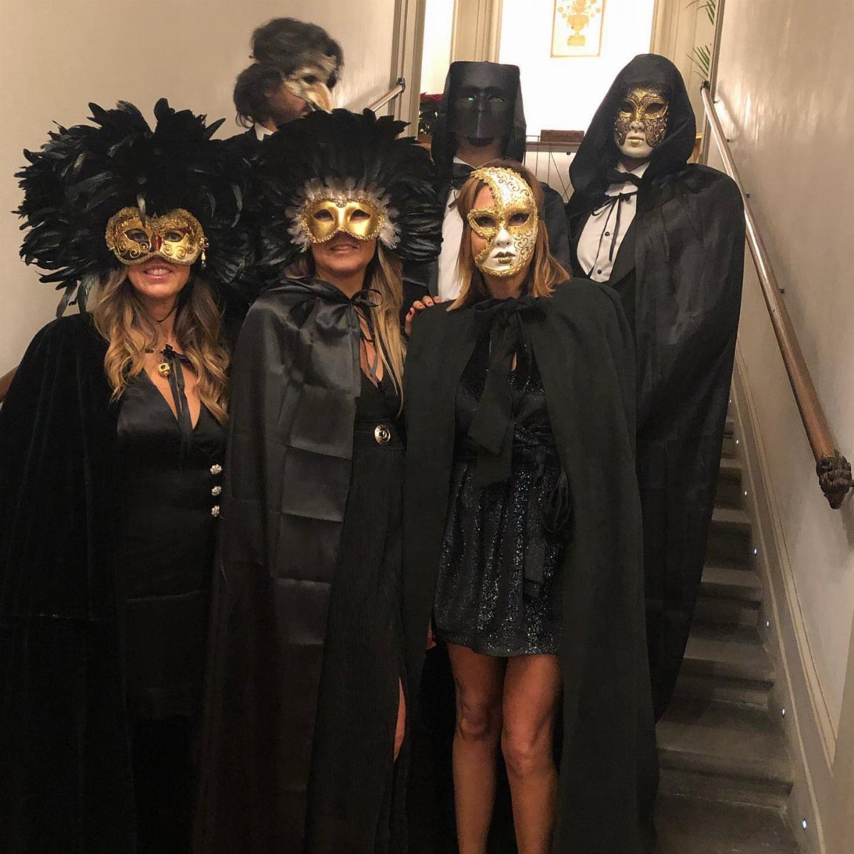 Adults in black formal capes and gold masquerade masks posing on a staircase.