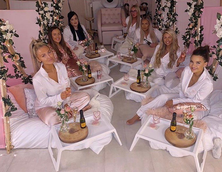 Women in satin robes relaxing on floor mattresses in a pink floral room.