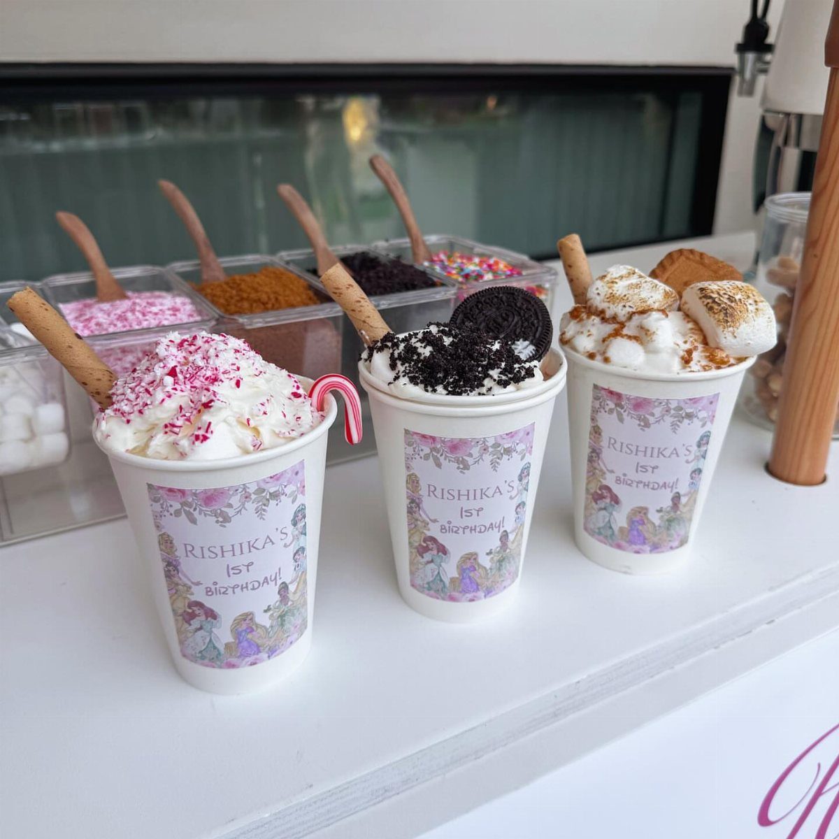 Three decorated hot cocoa cups with whipped cream and toppings at a birthday party.