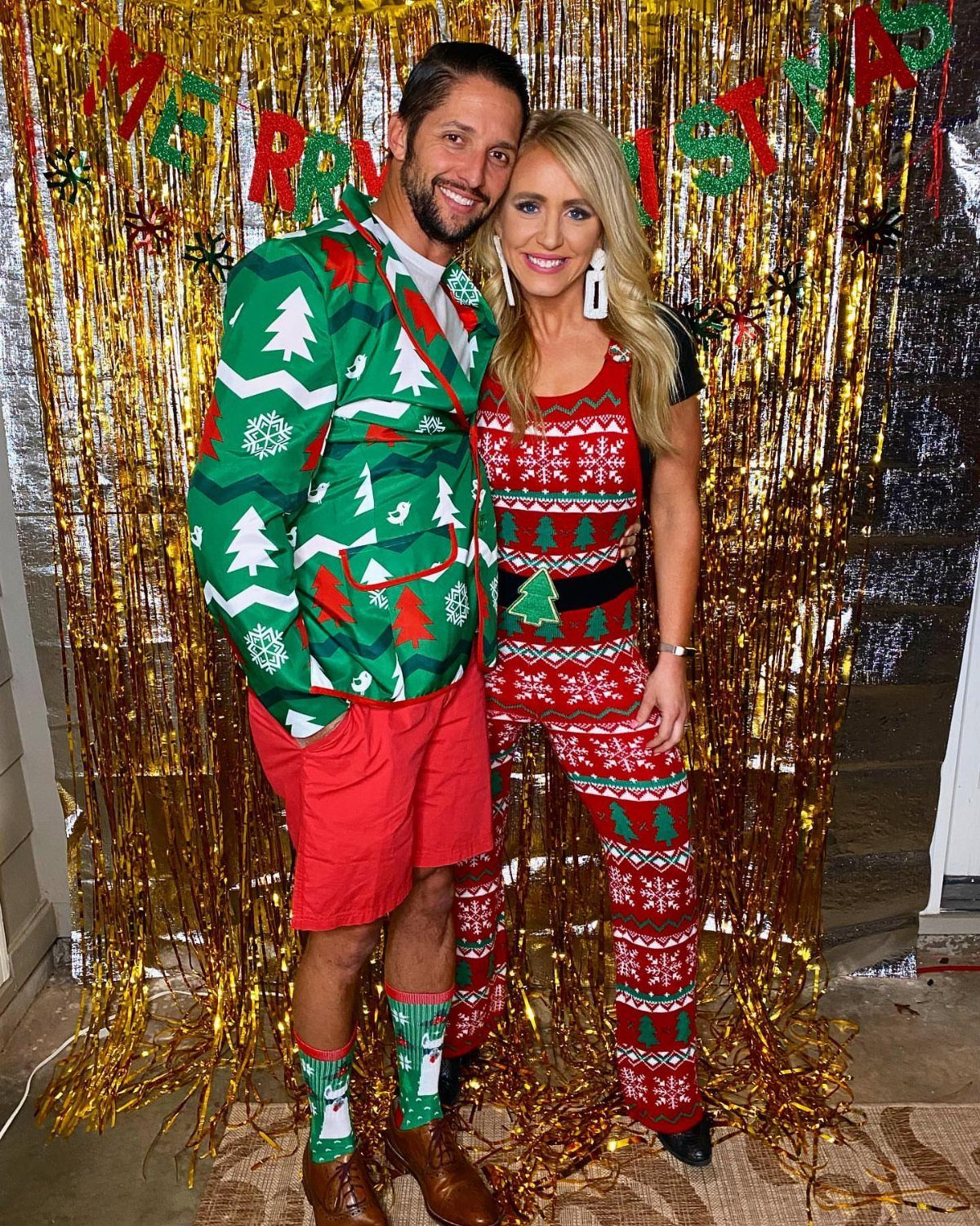 Man and woman wearing festive, quirky Christmas outfits posing before gold tinsel backdrop.