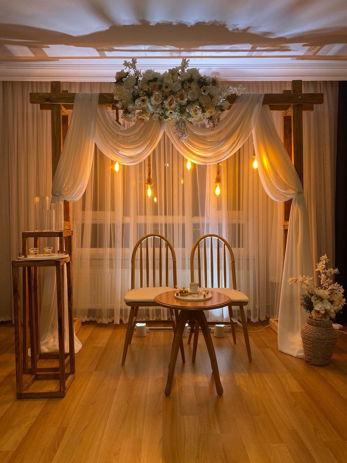 Wooden wedding arch with white floral arrangements and draped fabric over two chairs.