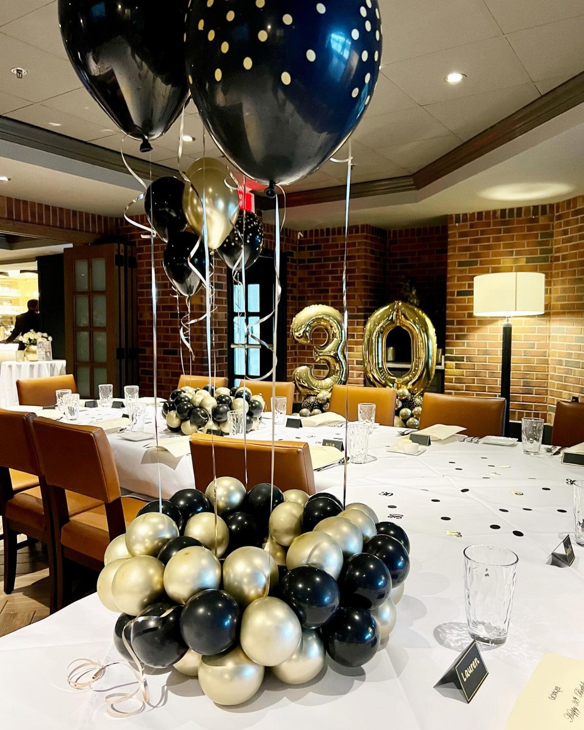Black and gold balloon centerpieces on white tables in a decorated banquet room.