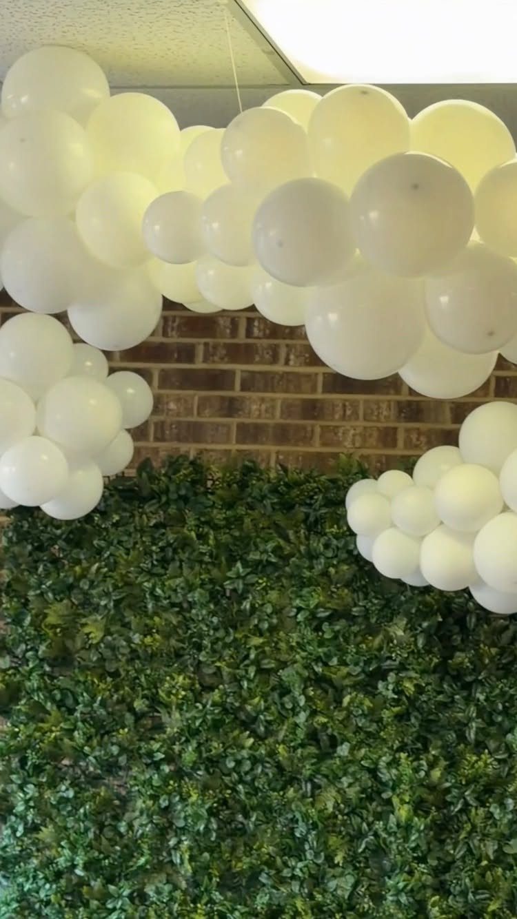 White balloon clouds hanging from a ceiling above a green hedge wall.