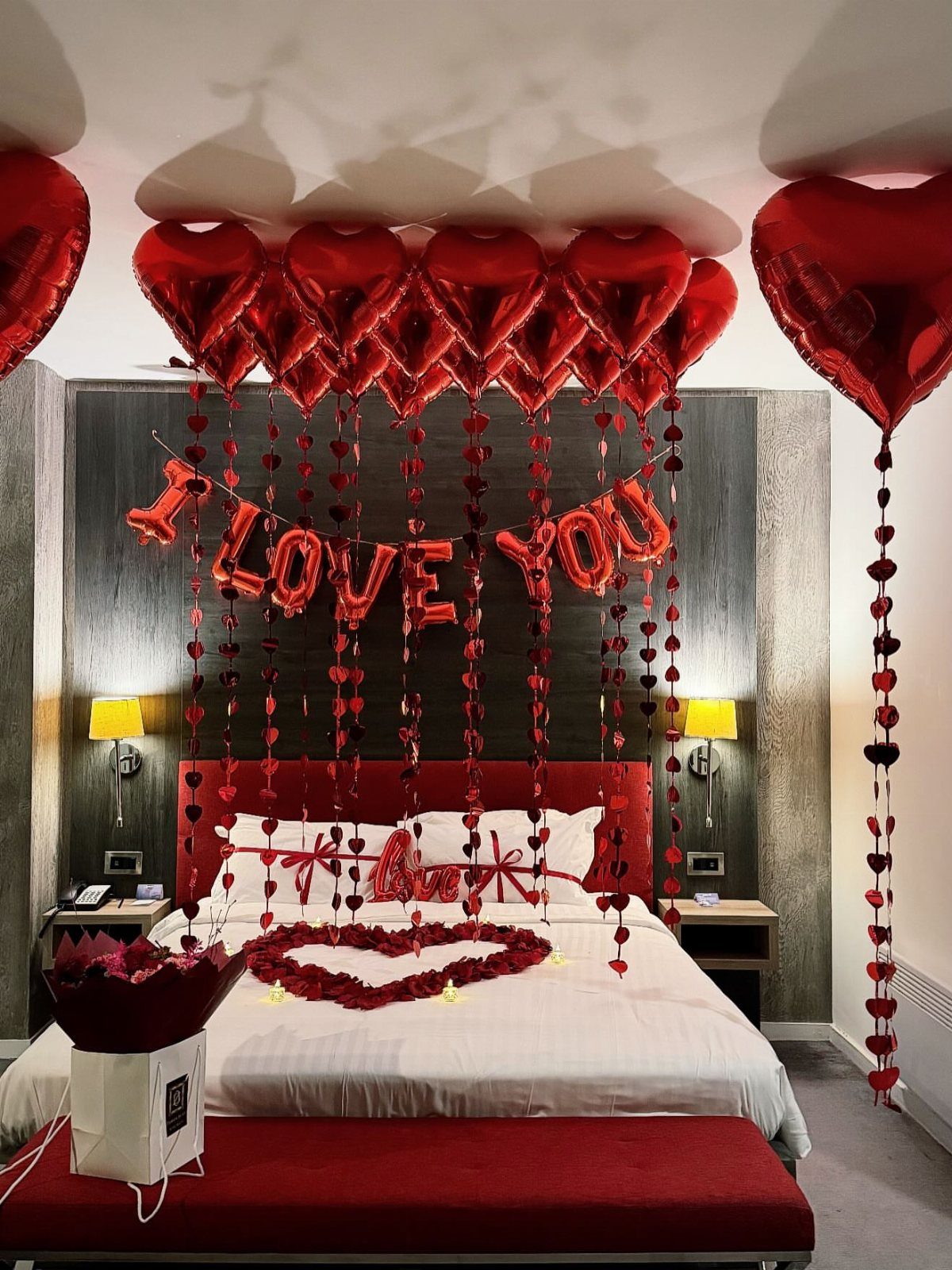 Red heart-shaped balloons floating on a bedroom ceiling above a decorated bed.