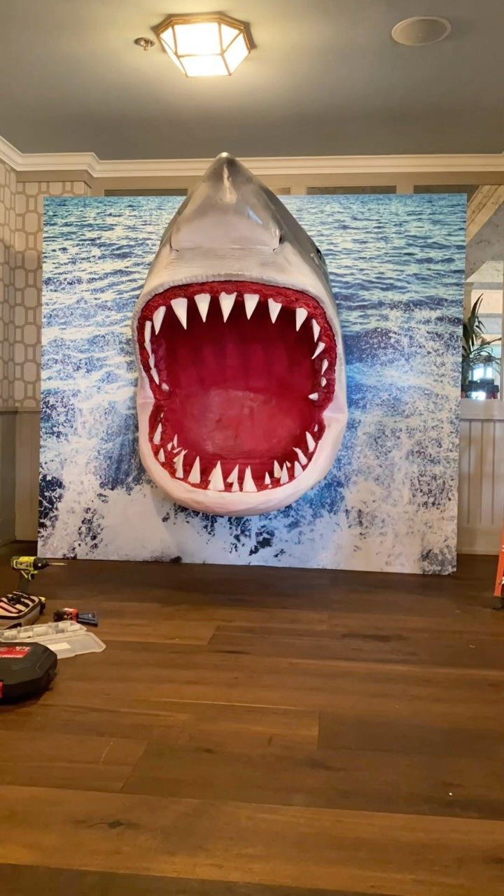 Large 3D shark head with open mouth against an ocean wave backdrop.