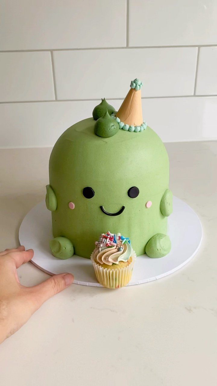Green dinosaur shaped cake wearing a party hat with a small sprinkle cupcake.