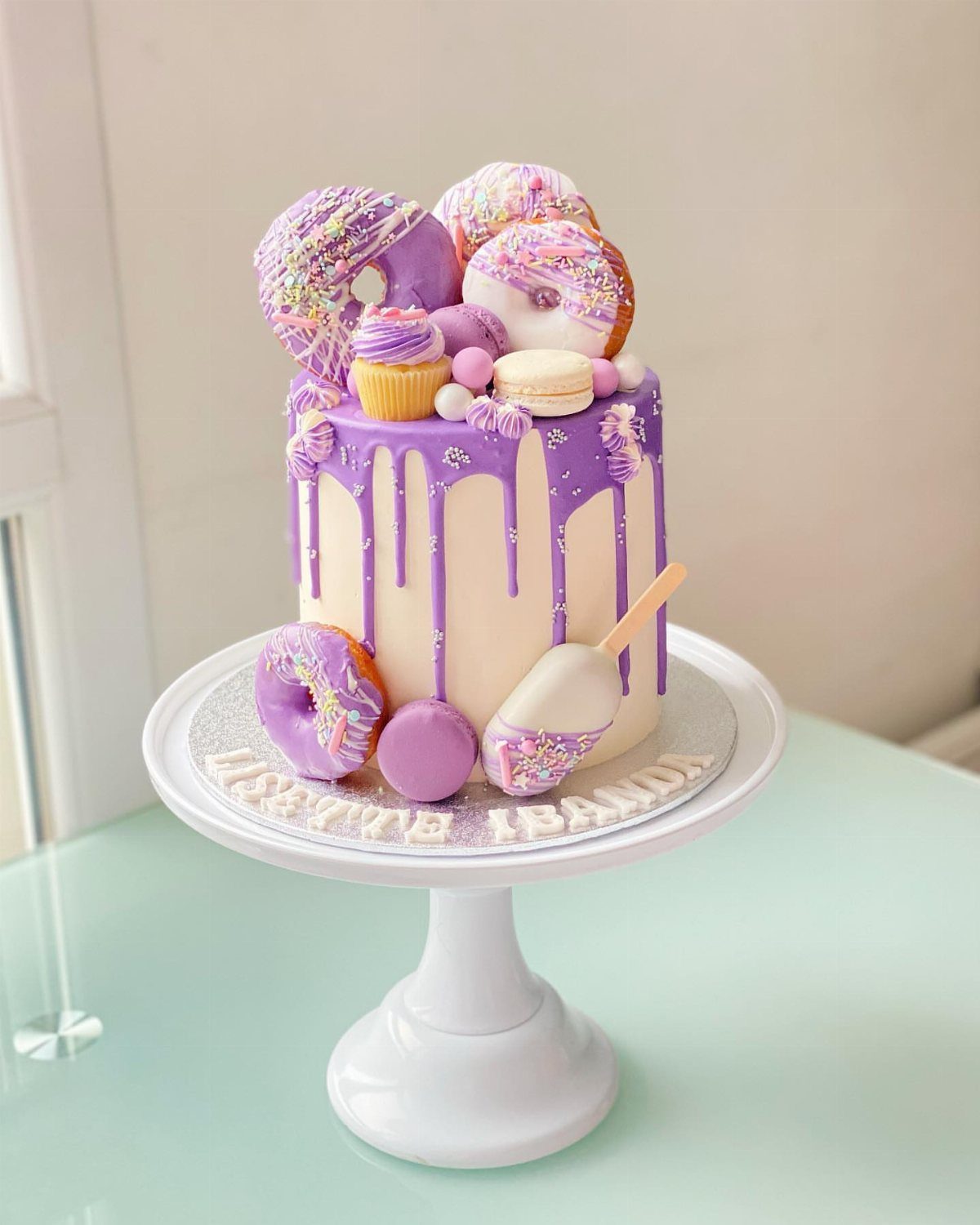 Purple drip cake topped with colorful donuts, macarons, and a cupcake on white stand