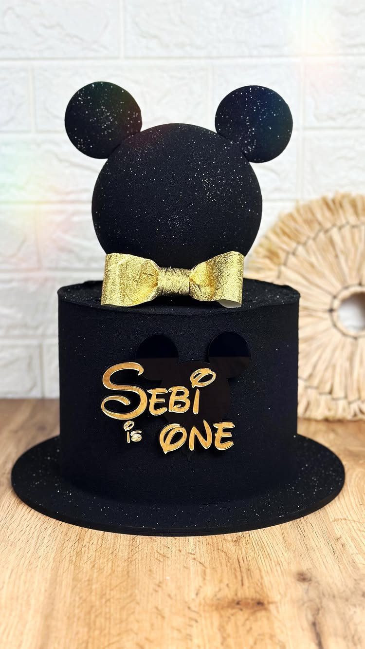 Black glittery Mickey Mouse themed cake with gold bow and 'SEBI IS ONE' text.