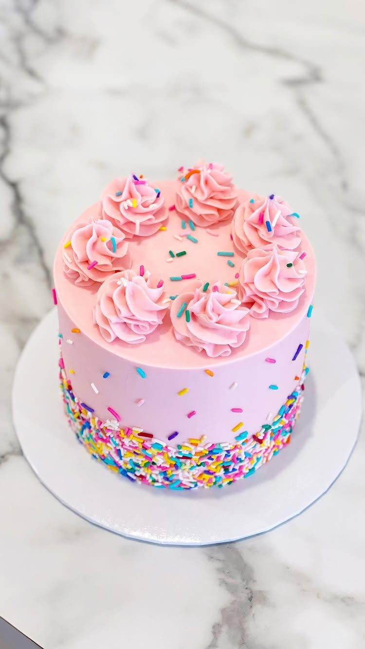Pink frosted cake with colorful sprinkles around the base and swirls on top.