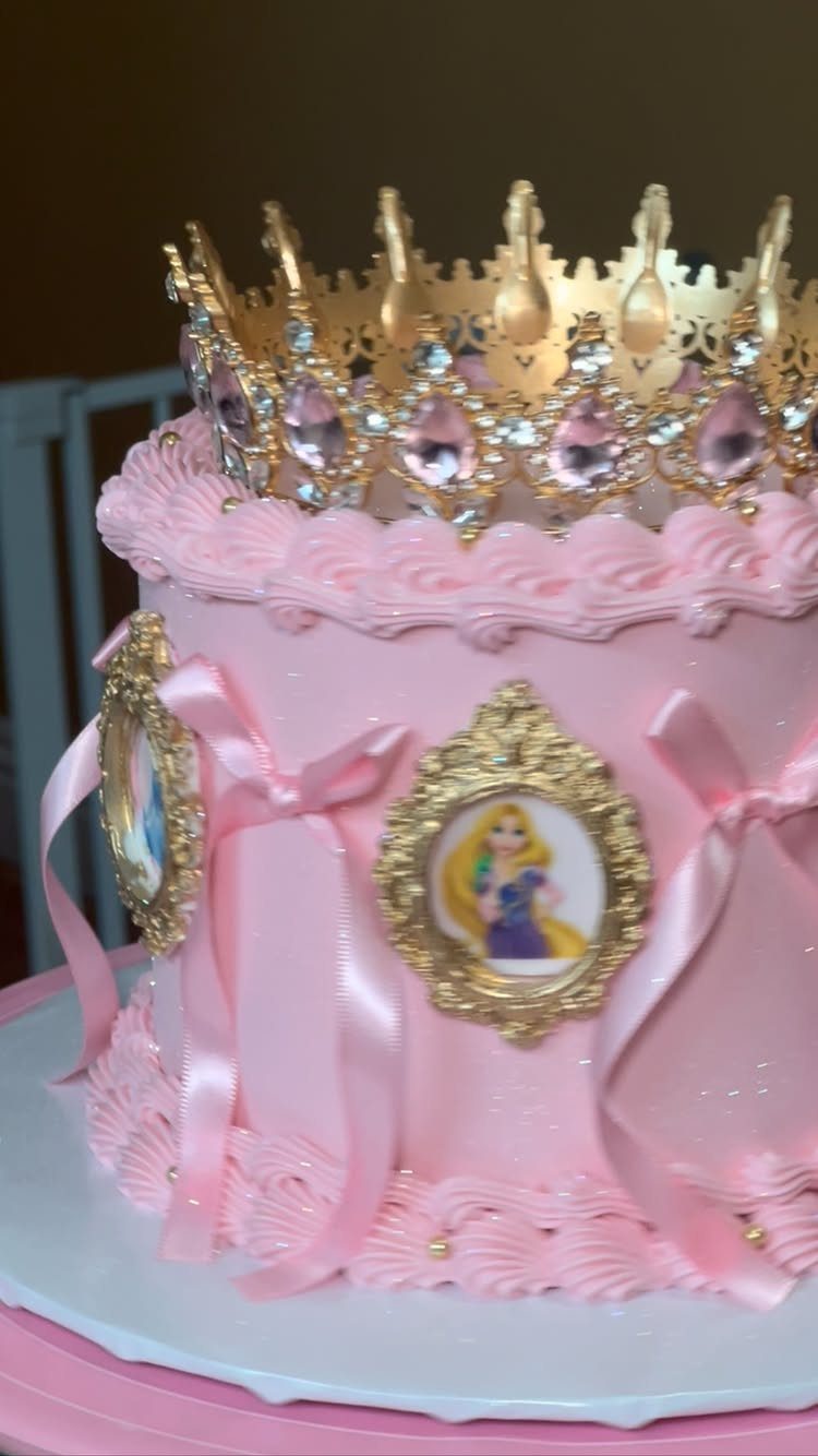 Pink princess cake with gold crown, ribbons, and princess portraits on a stand