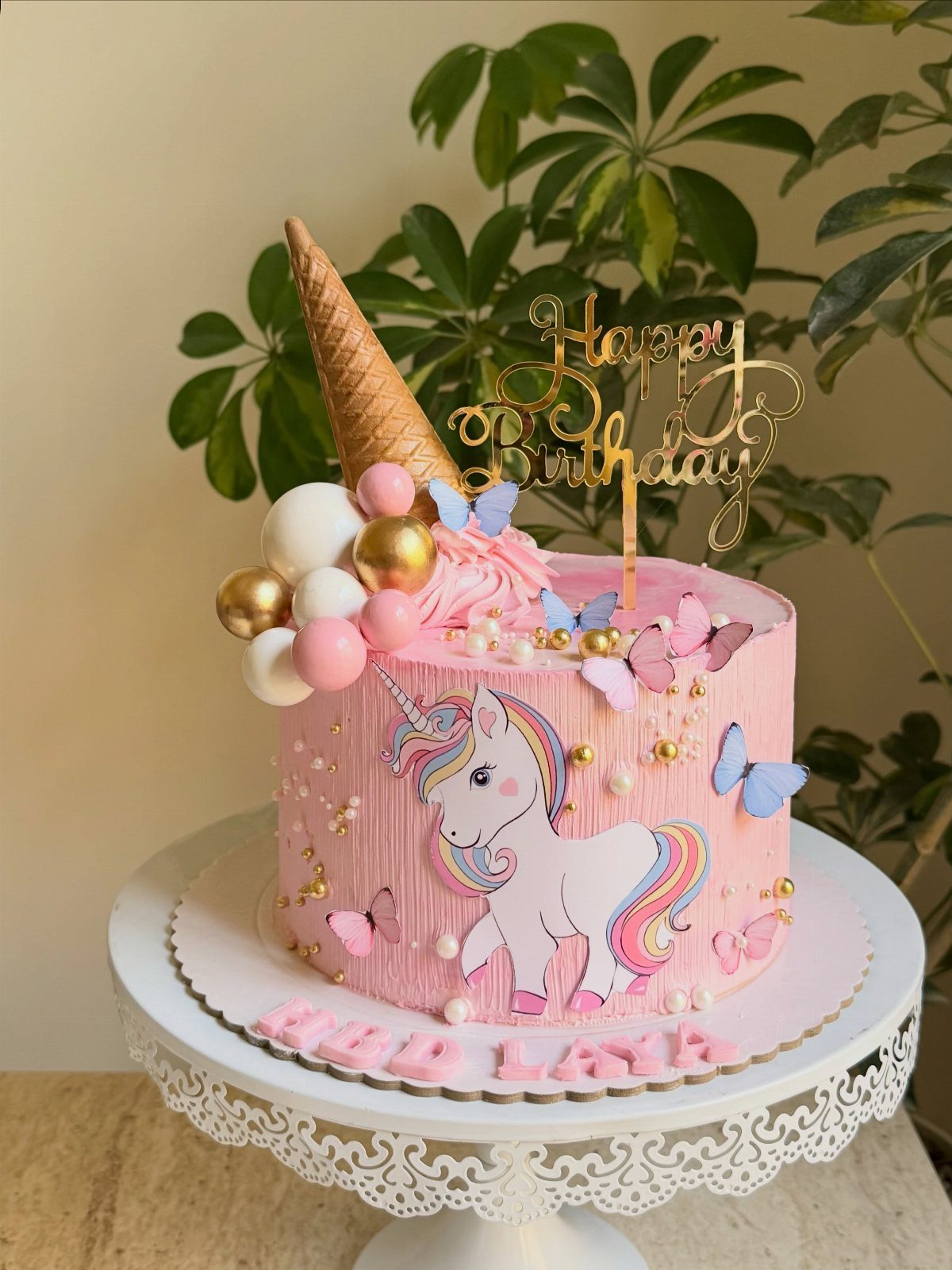 Pink unicorn birthday cake with butterflies, gold pearls, and an ice cream cone topper.