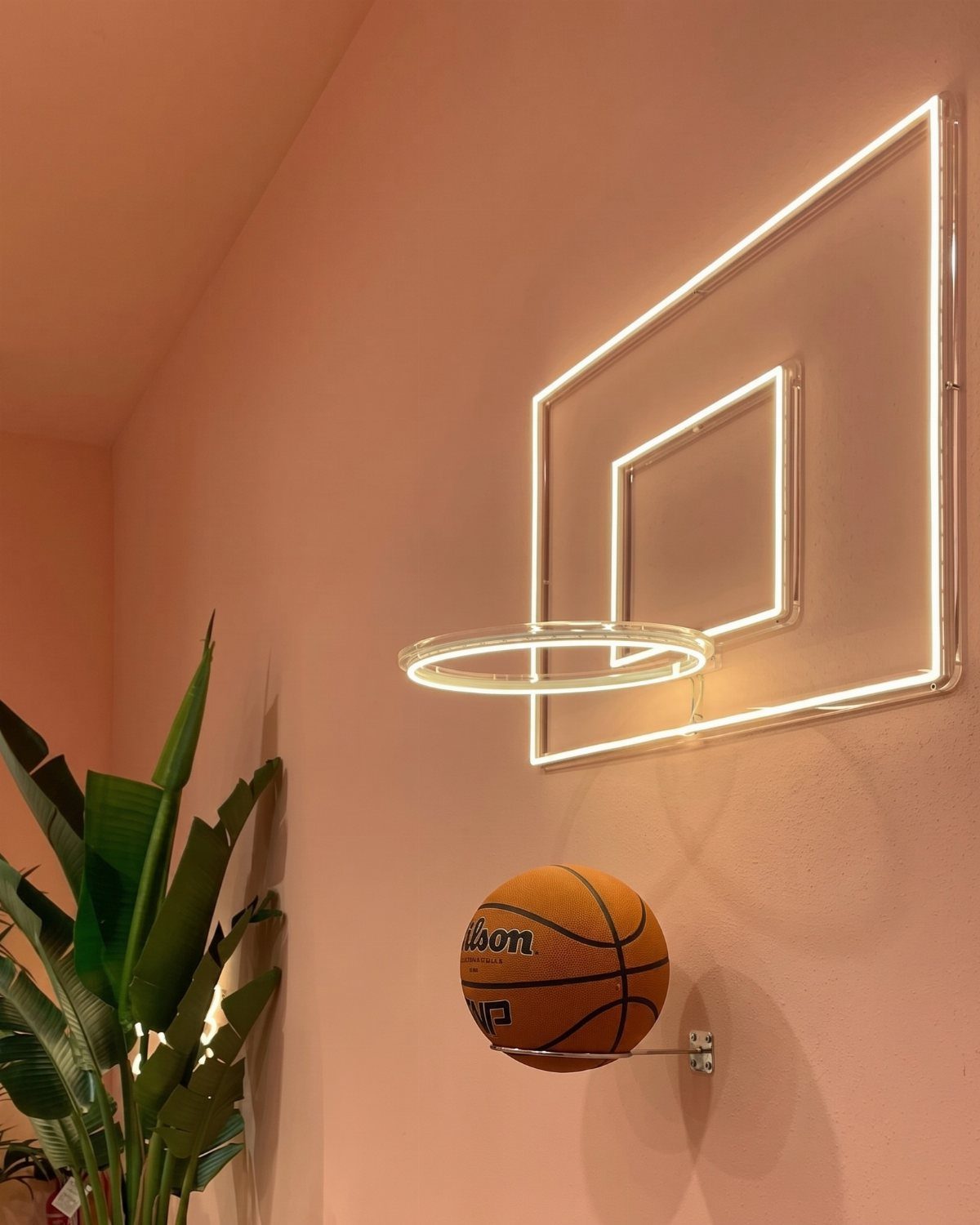 Neon basketball hoop light fixture on a peach wall with a basketball below.