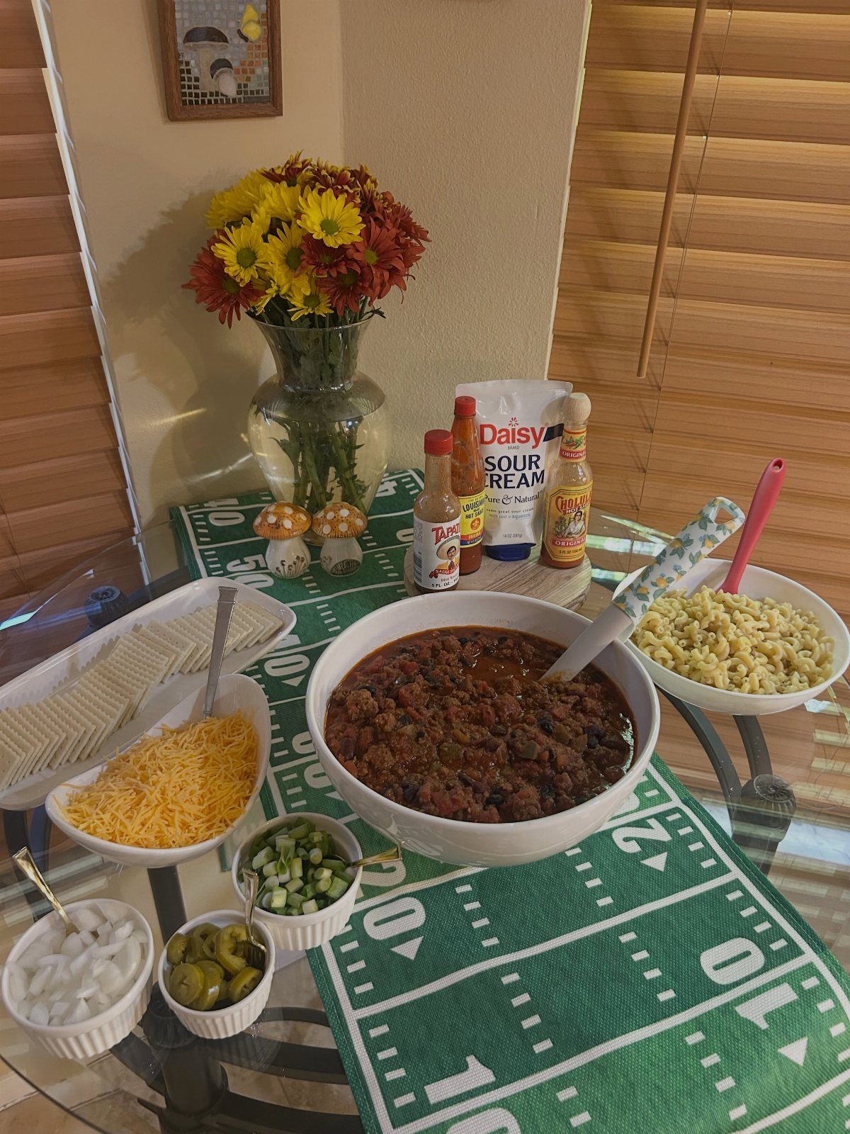 Chili bar setup on a football field table runner with various toppings and flowers.