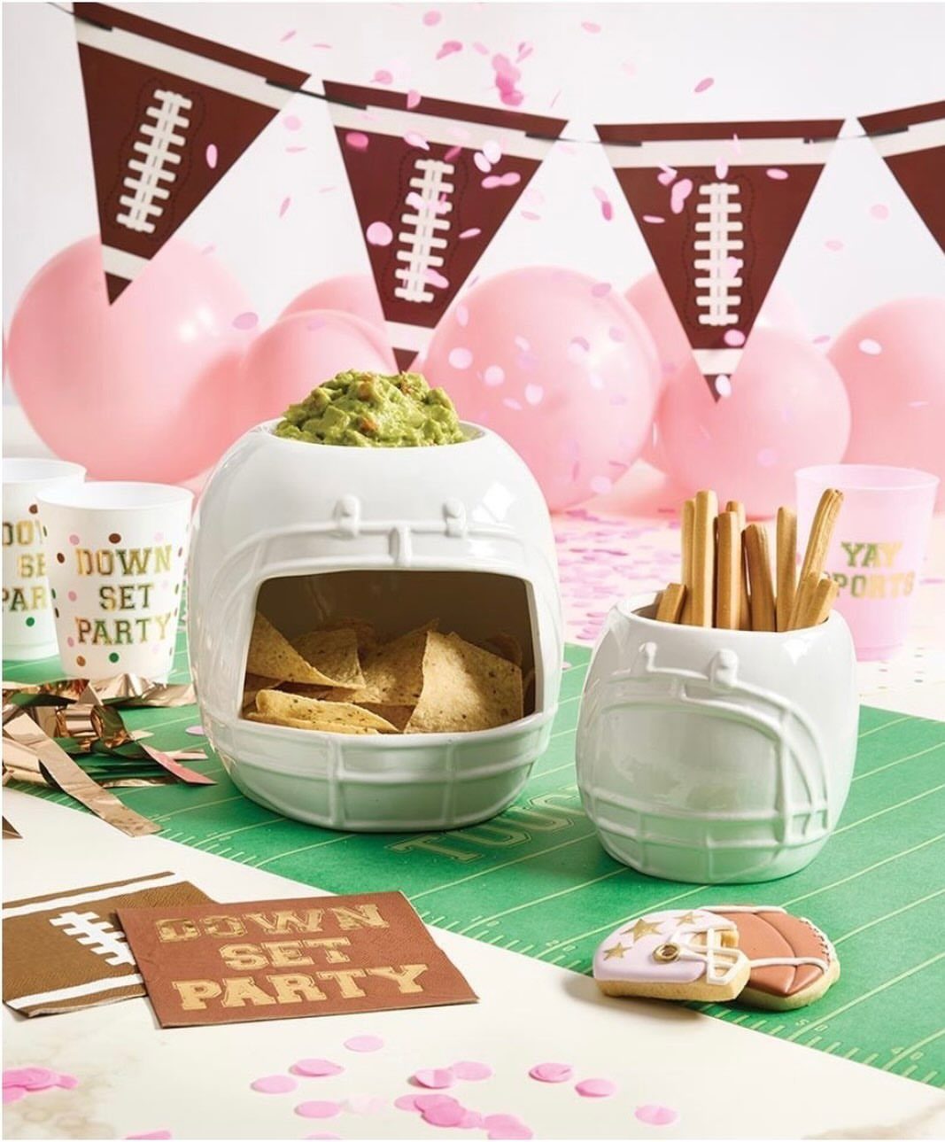 Football-shaped ceramic bowls with snacks on a green runner with pink balloons.