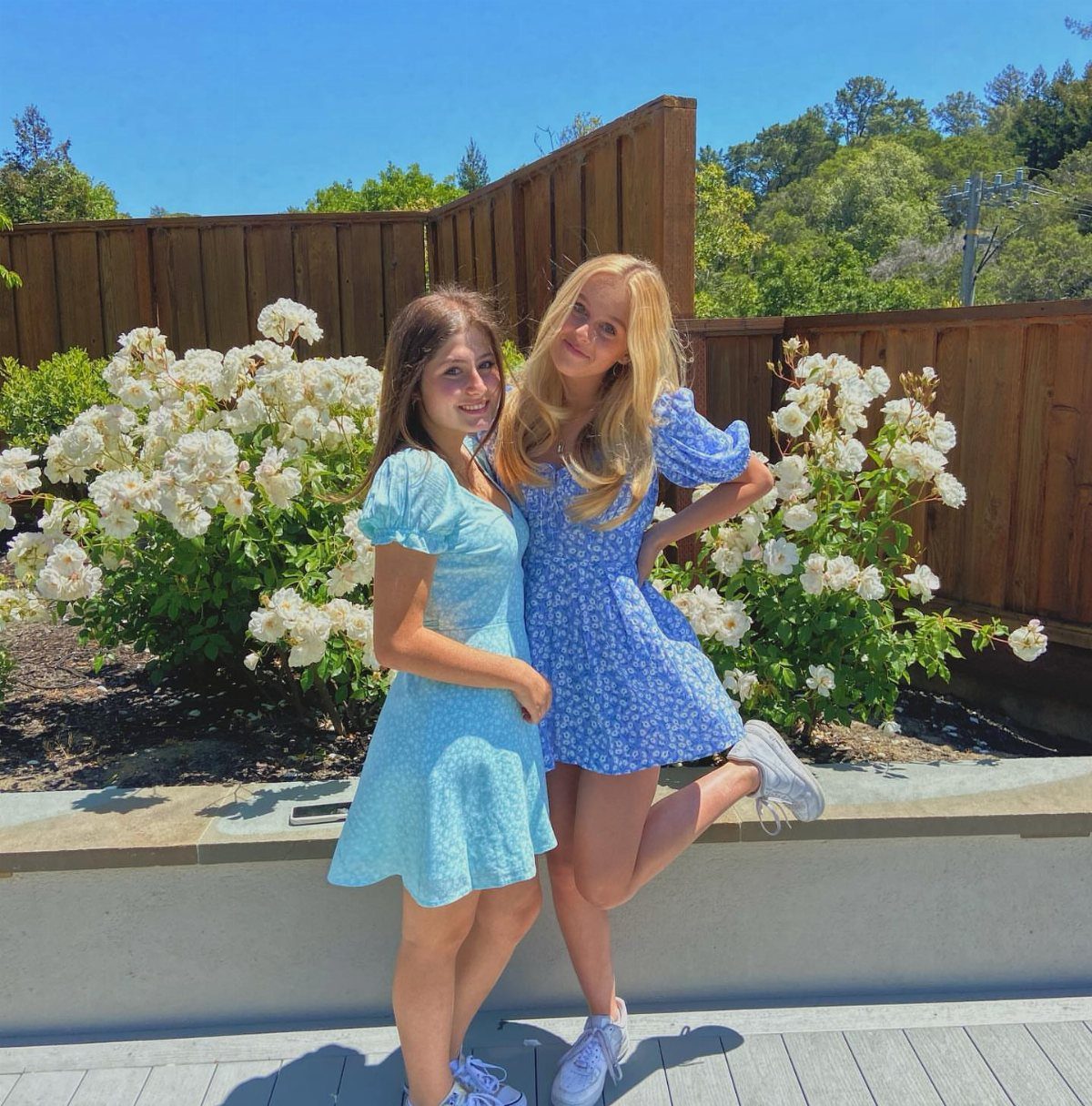 Two teenage girls wearing short light blue floral dresses and white sneakers outdoors.