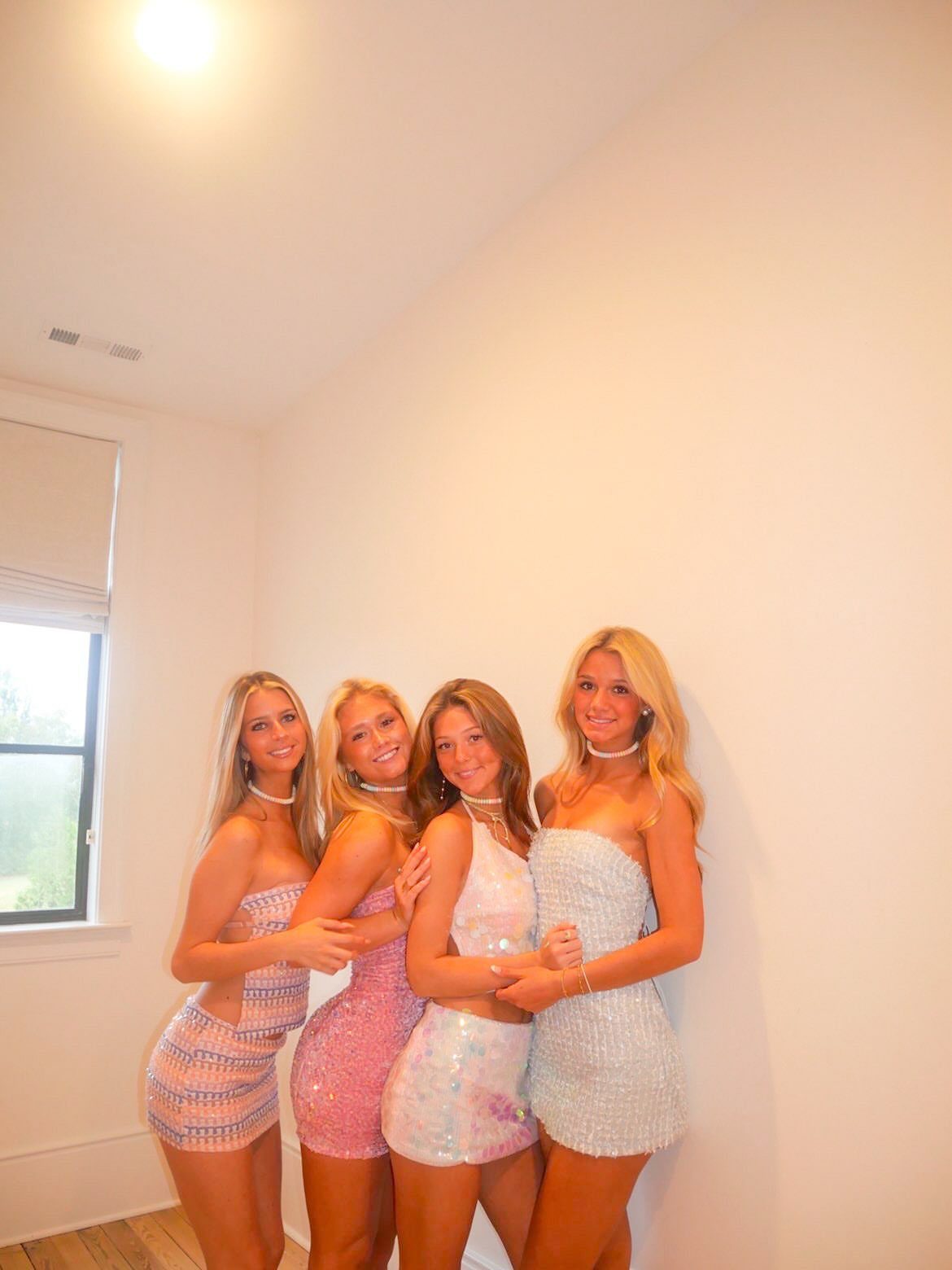 Four young women wearing sparkly pastel sequin dresses posing against a white wall.