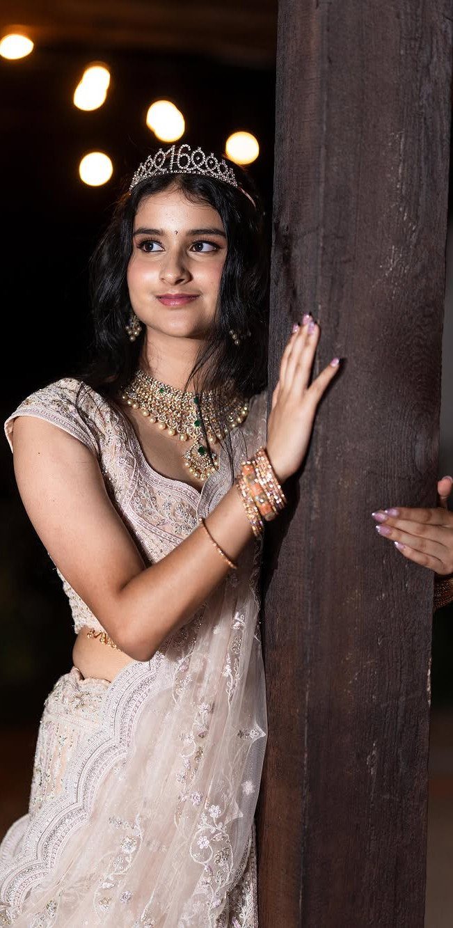 Young woman in a pink embroidered lehenga and tiara leaning against a wooden pillar.