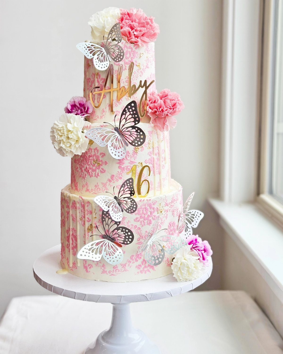 Three-tier white cake with pink floral patterns, buttercream flowers, and decorative butterflies.