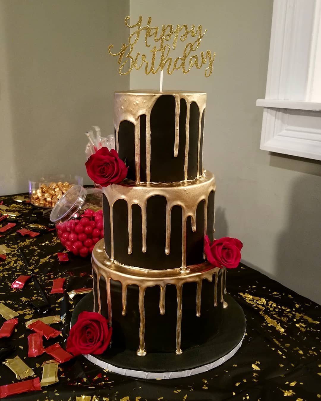 Three-tier black cake with gold drips, red roses, and a Happy Birthday topper.