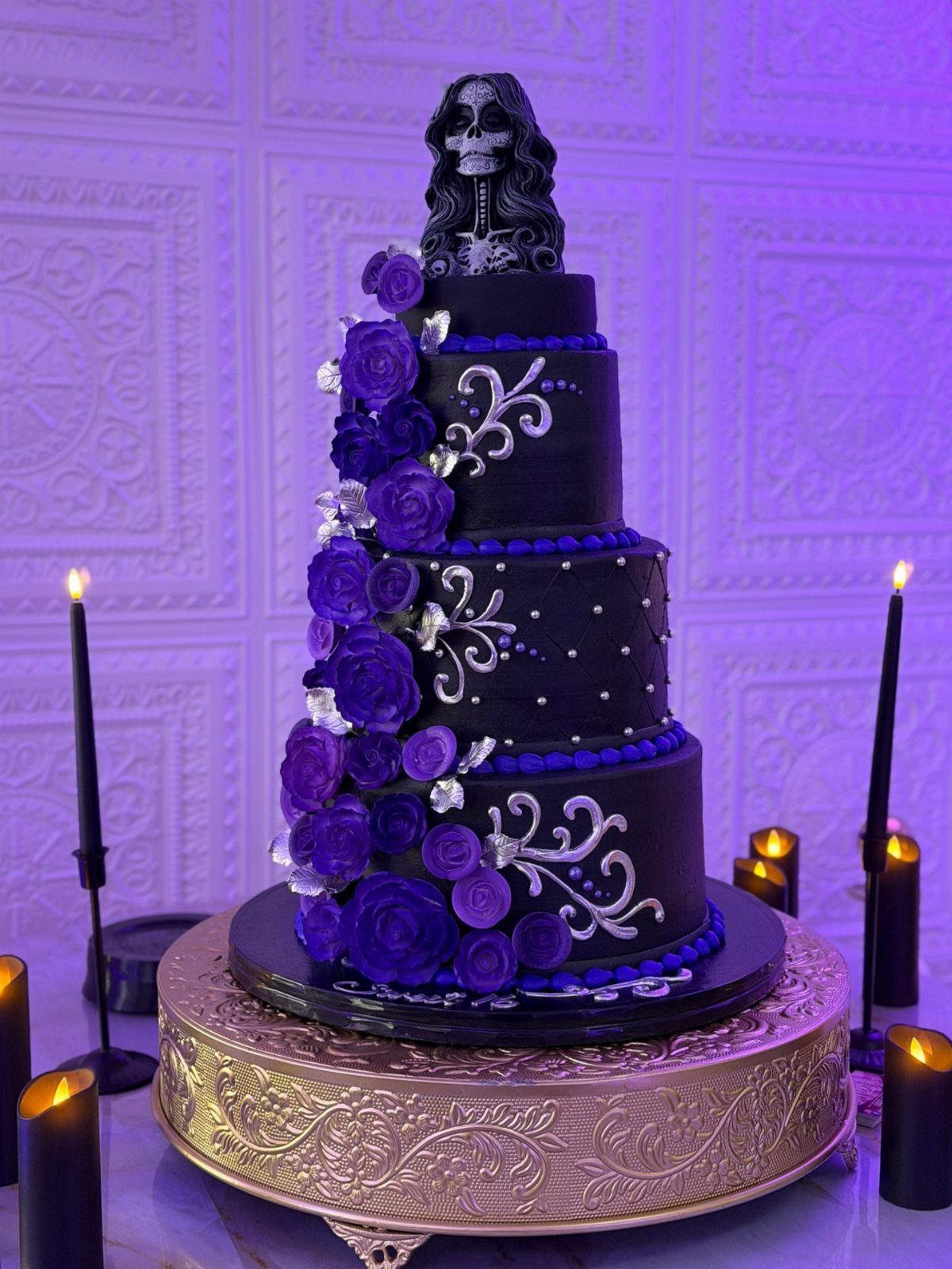 Four-tier black cake with purple roses and a skeleton topper on gold stand.