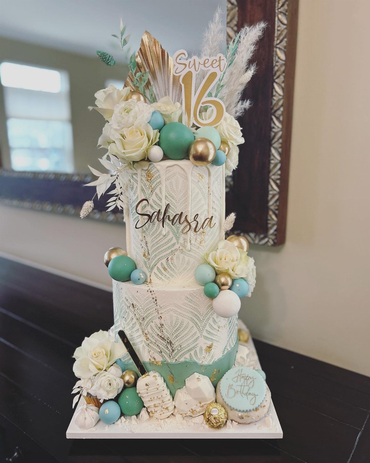 Two-tier white sweet 16 cake with teal spheres, white roses, and gold accents.