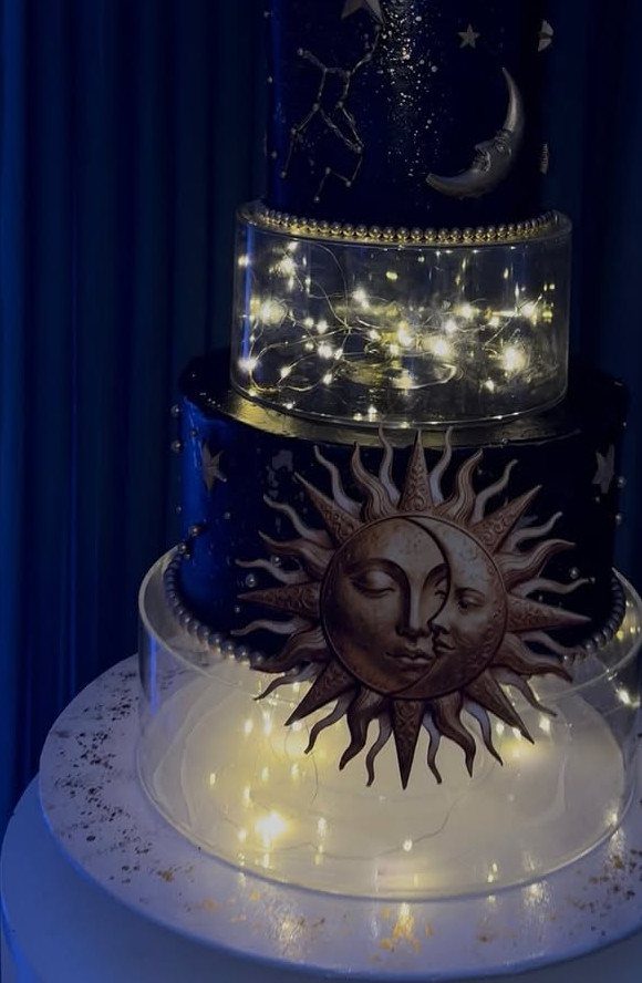 Dark blue celestial cake with gold moon and sun motifs and glowing fairy lights.