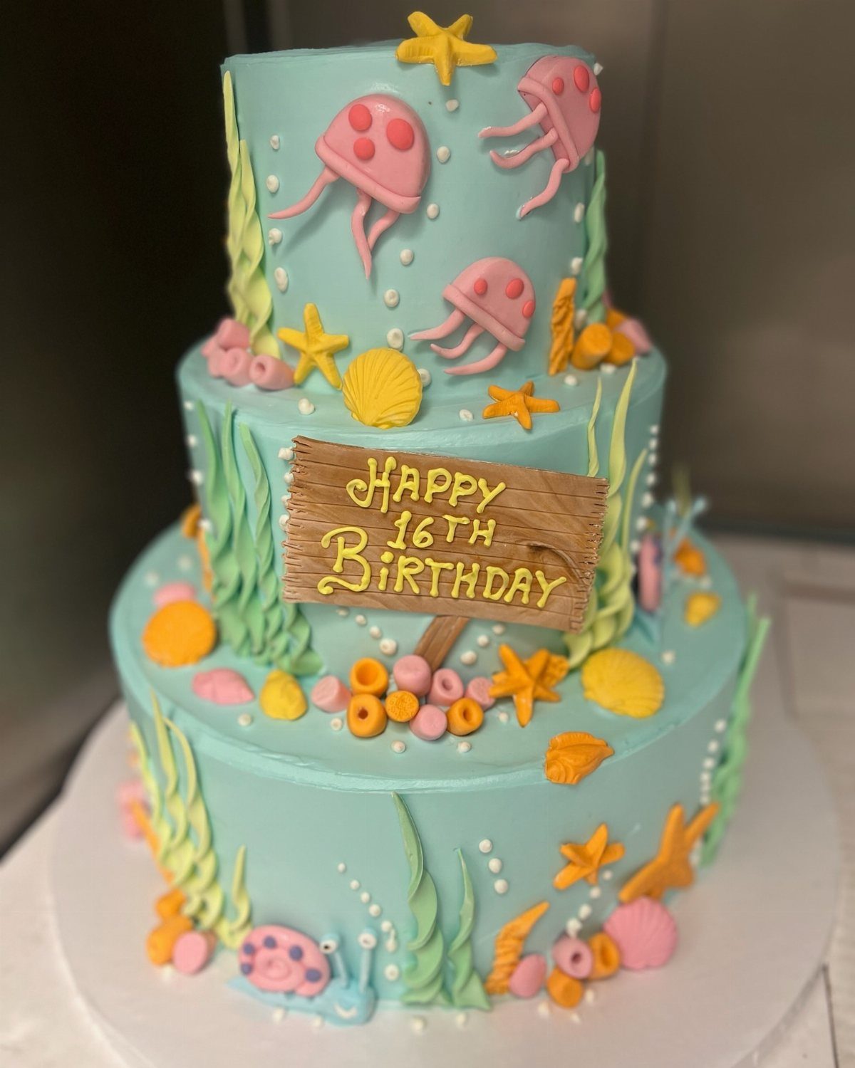 Three-tier blue birthday cake decorated with pink jellyfish, yellow starfish, and seashells.