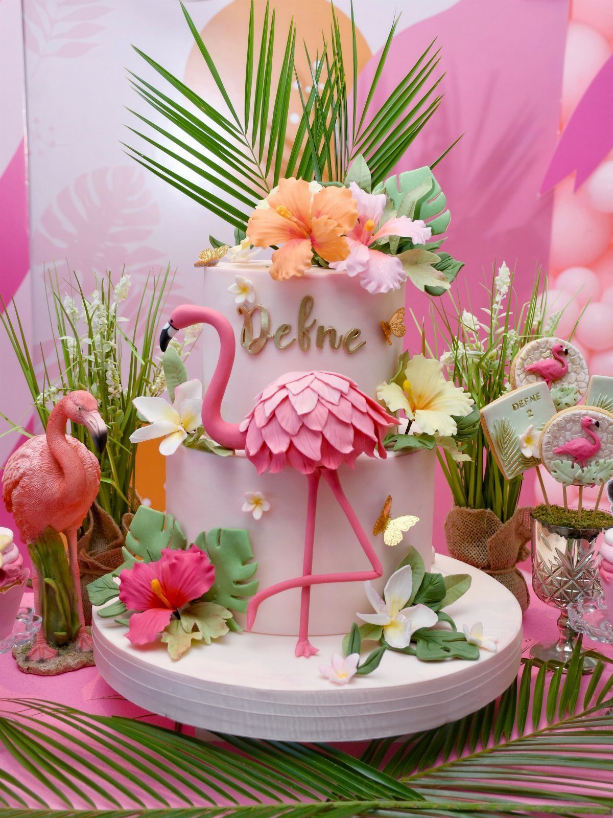 Two-tier white cake with a pink flamingo, tropical flowers, and palm leaves.