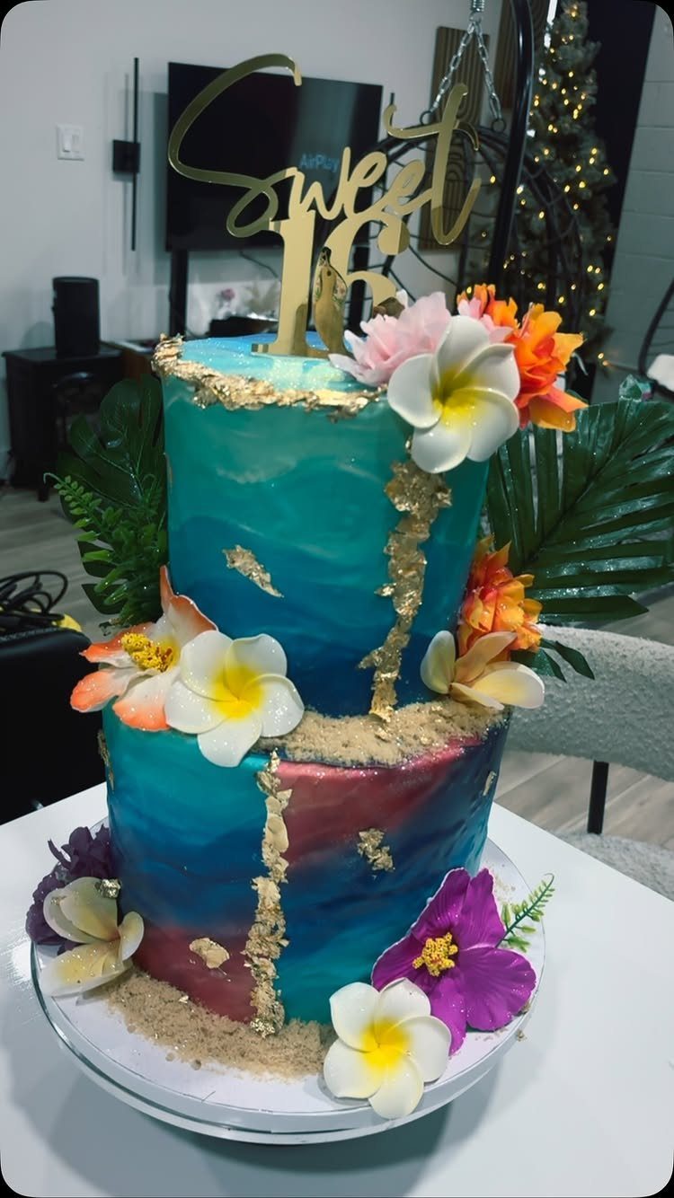 Two-tier watercolor cake with blue and magenta frosting, gold accents, and tropical flowers.