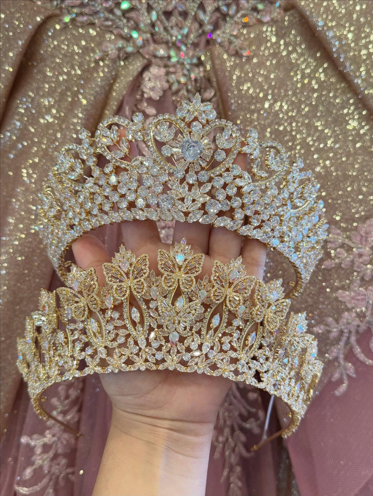 Two sparkling crystal tiaras held in front of a shimmering rose-gold sequined gown.