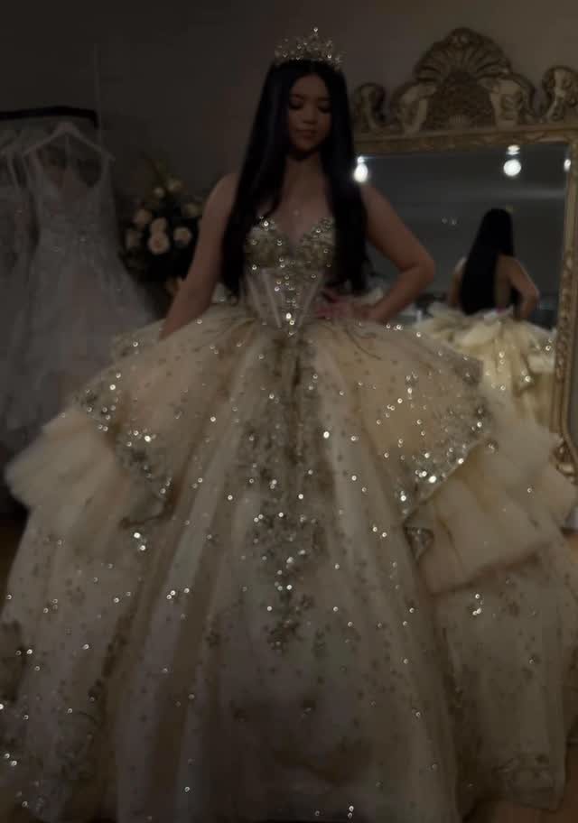 Young woman in a sparkling champagne ball gown and tiara standing before a mirror