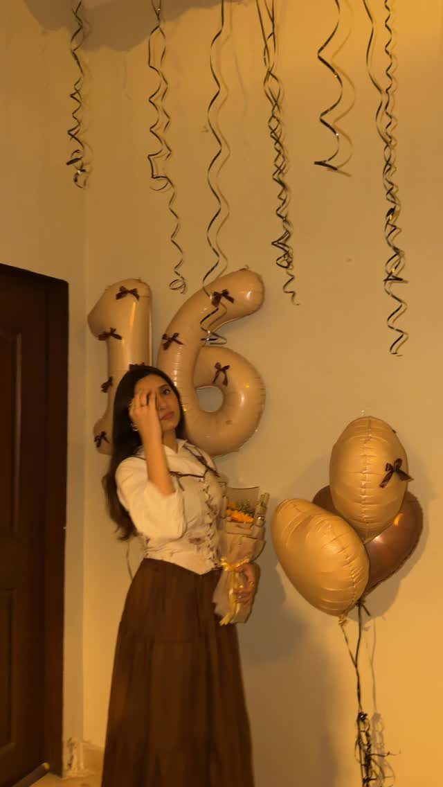 Young woman in white blouse and brown skirt posing with gold 16 balloons.