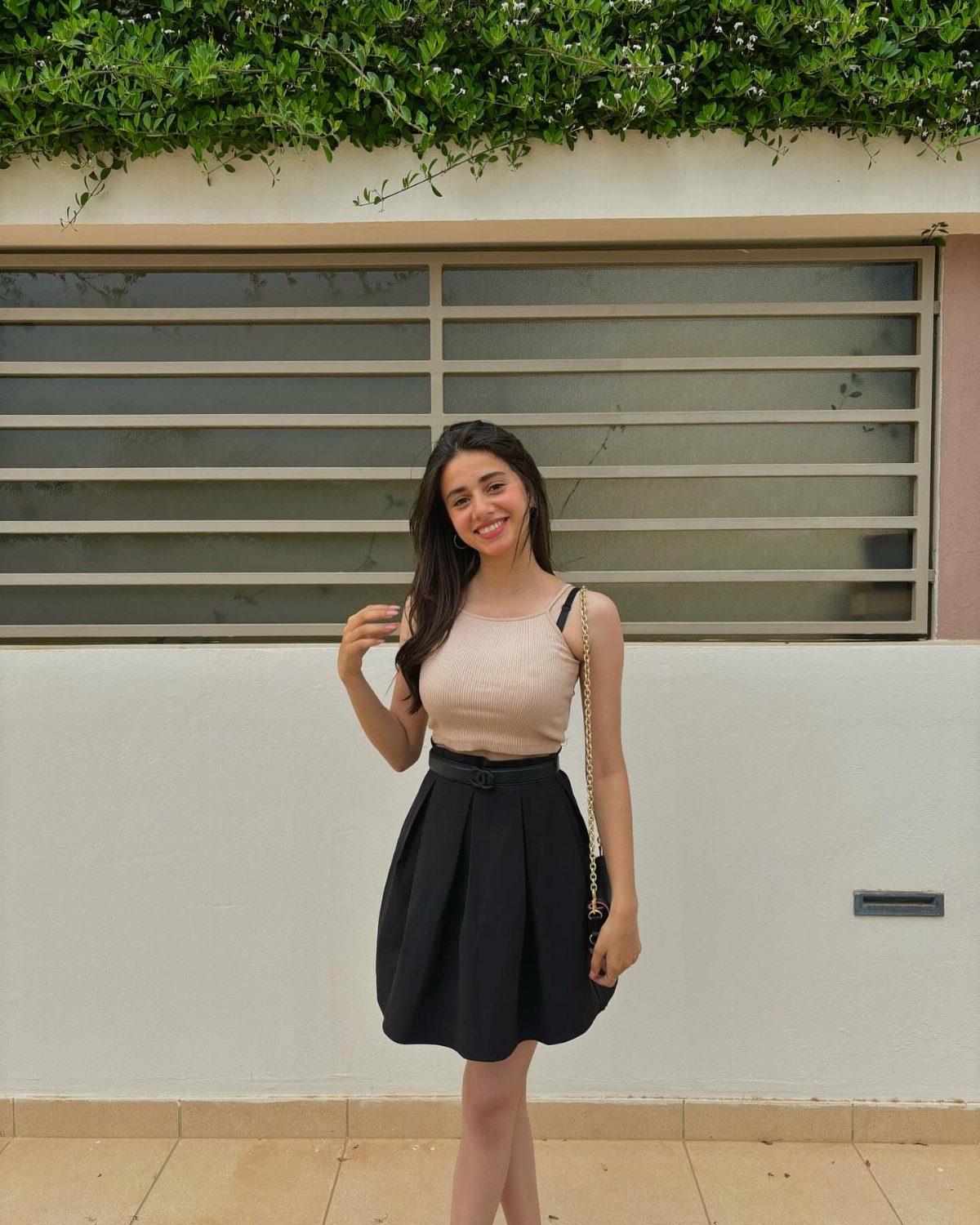 Young woman wearing a beige top and short black pleated skirt posing outdoors.