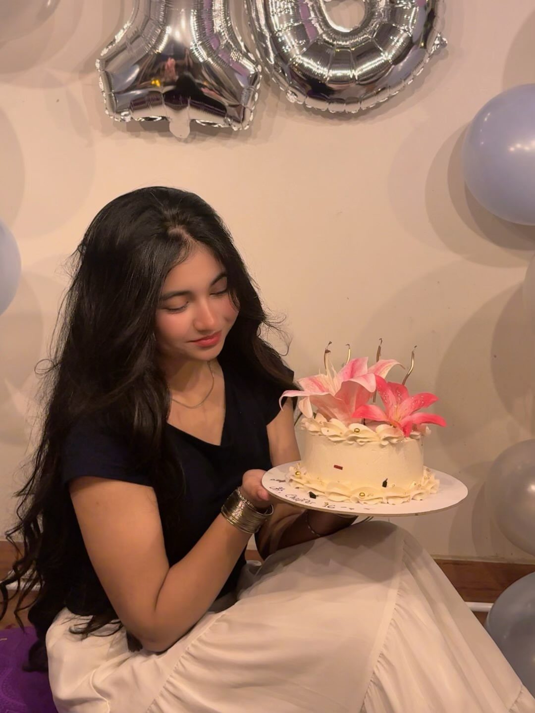Young woman in navy top and white skirt holding a lily-decorated cake.